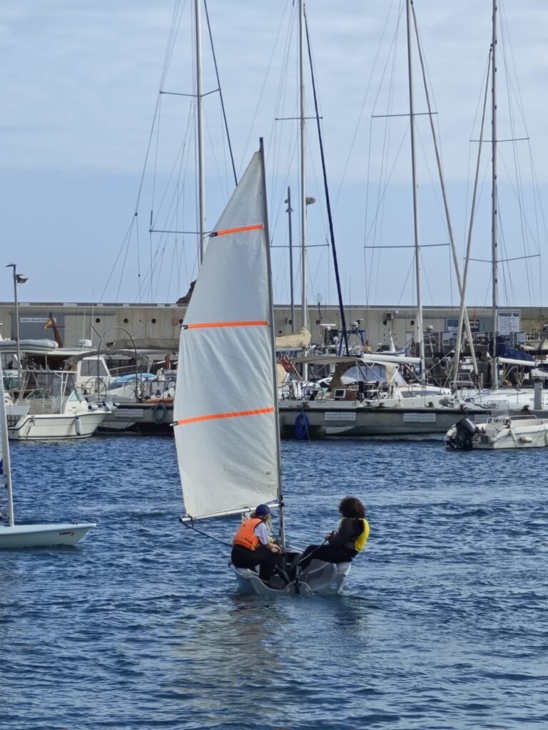 Dos jóvenes navegantes disfrutan de un día soleado en el mar, aprendiendo a controlar su velero.